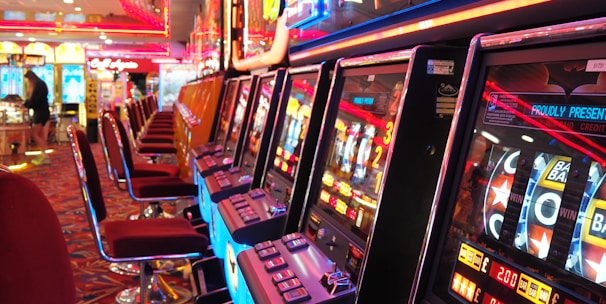 A brightly lit casino interior featuring a row of colorful slot machines. The neon lights create a vibrant atmosphere, with patterned carpeting adding to the decor. Stools are aligned neatly in front of each machine.