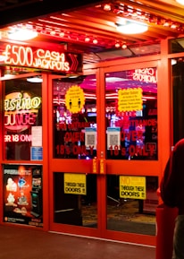 A brightly lit entrance to a casino with neon signs, including one advertising a £500 jackpot. The doors have clear signage indicating entry is restricted to those over 18. A person in a red jacket stands to the right, possibly interacting with a machine or display.