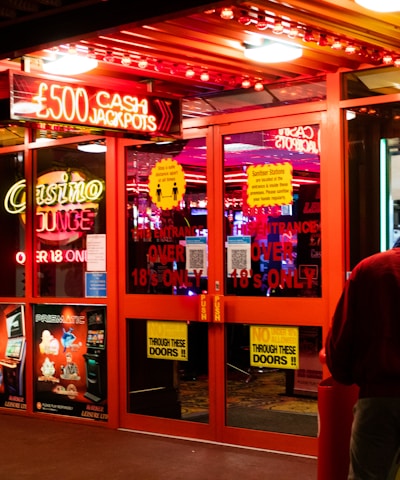 A brightly lit entrance to a casino with neon signs, including one advertising a £500 jackpot. The doors have clear signage indicating entry is restricted to those over 18. A person in a red jacket stands to the right, possibly interacting with a machine or display.