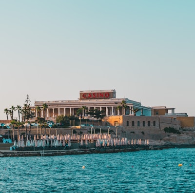 A seaside view of a large building labeled as a casino, situated on a rocky shore. The building is surrounded by palm trees and other smaller structures. The calm sea water reflects the sky, and there are several closed umbrellas lined along the waterfront.